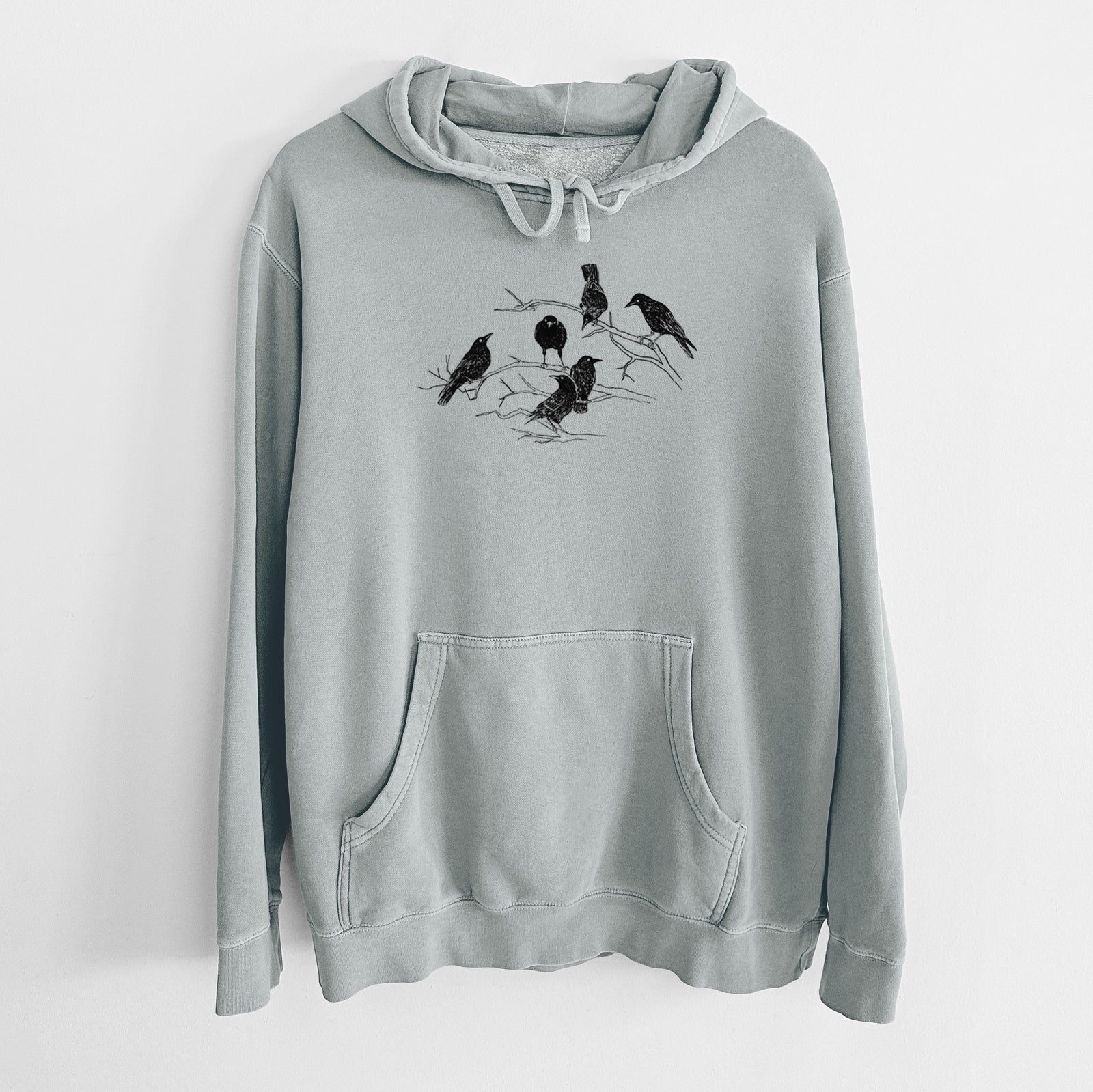 Sage green pigment dyed hoodie laid flat, printed with a black ink illustration of a group of crows perched on bare branches.