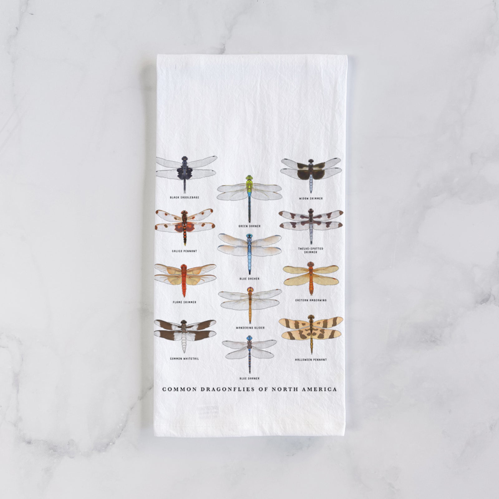 White cotton tea towel printed with a full-color chart of North American dragonflies, each labeled with its species, on a marble counter beside a round wooden cutting board and greenery.