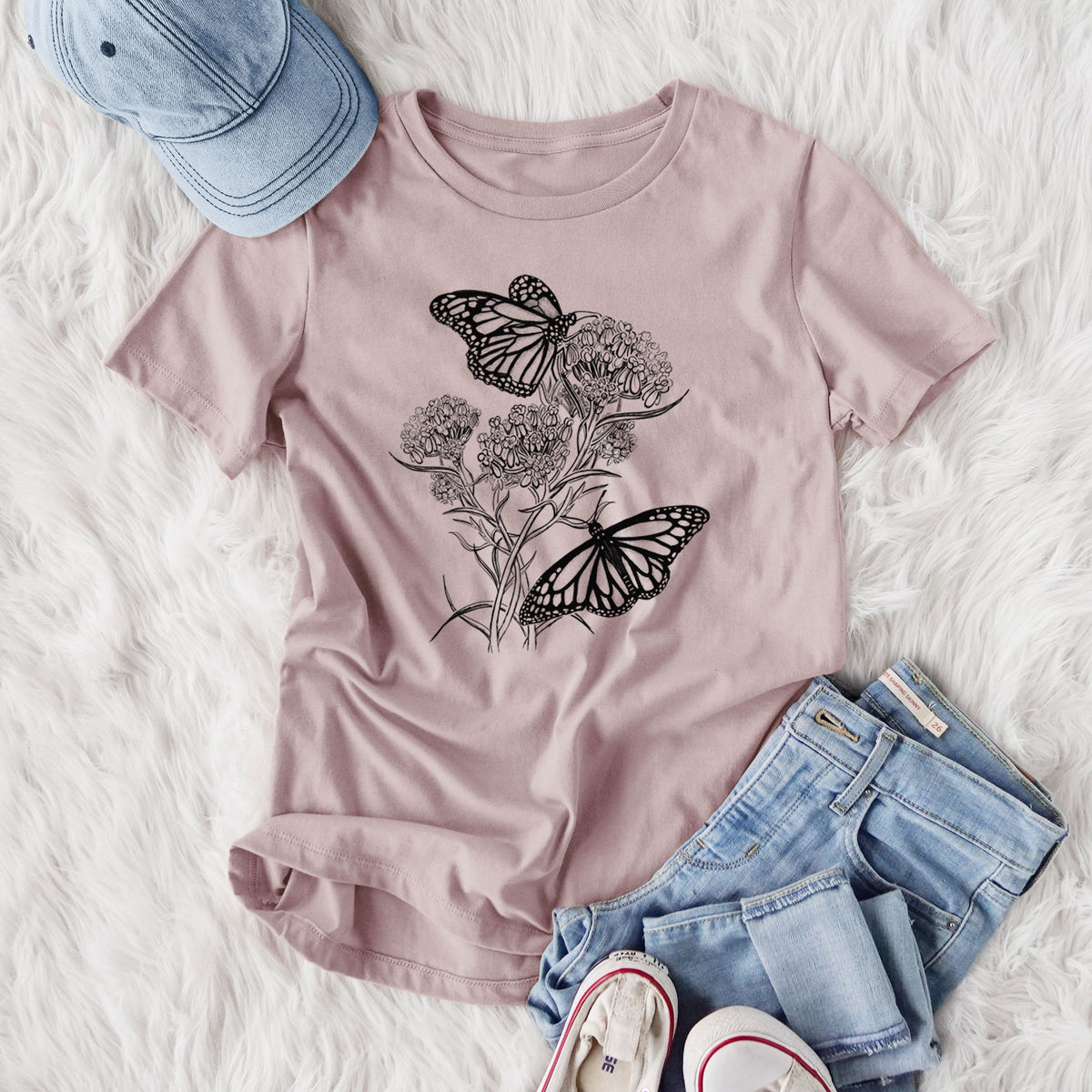 Women's relaxed fit cotton t-shirt printed with a black ink illustration of two monarch butterflies on flowering milkweed, styled flat on a white faux fur rug with jeans.