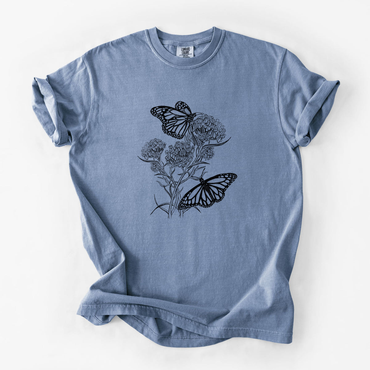 Blue jean Comfort Colors t-shirt laid flat, printed with a black ink illustration of two monarch butterflies on flowering milkweed.