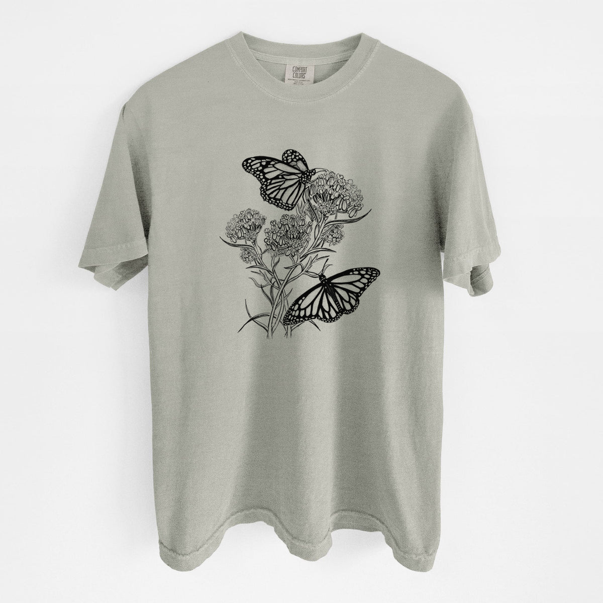 Sandstone beige Comfort Colors t-shirt laid flat, printed with a black ink illustration of two monarch butterflies on flowering milkweed.