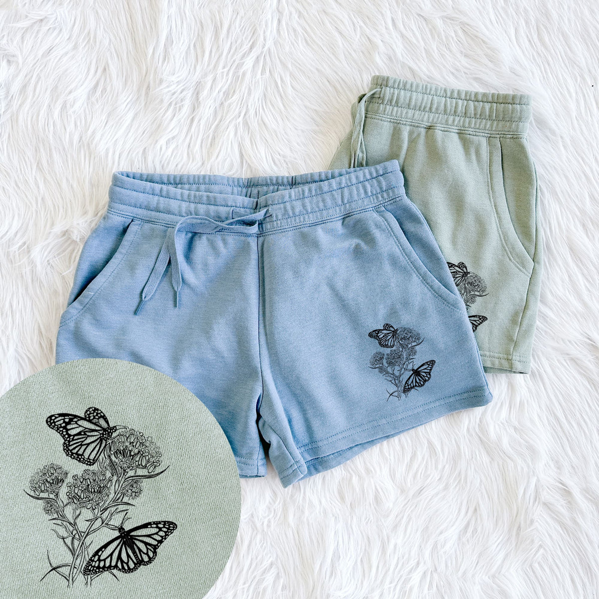 Two pairs of Cali Wave shorts in different colors printed with a black ink illustration of two monarch butterflies on flowering milkweed, with a close-up of the print.