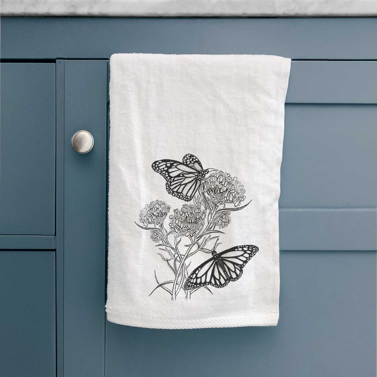 White hand towel printed with a black ink illustration of two monarch butterflies on flowering milkweed, hanging on a cabinet door.