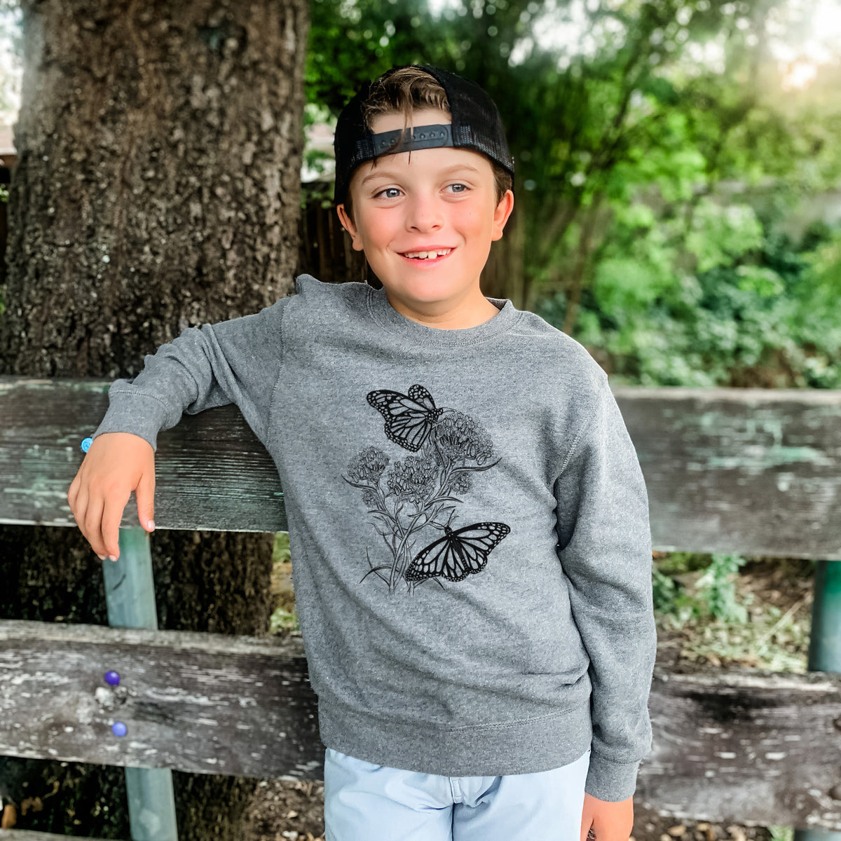 Boy wearing a youth crewneck sweatshirt printed with a black ink illustration of two monarch butterflies on flowering milkweed, outdoors.