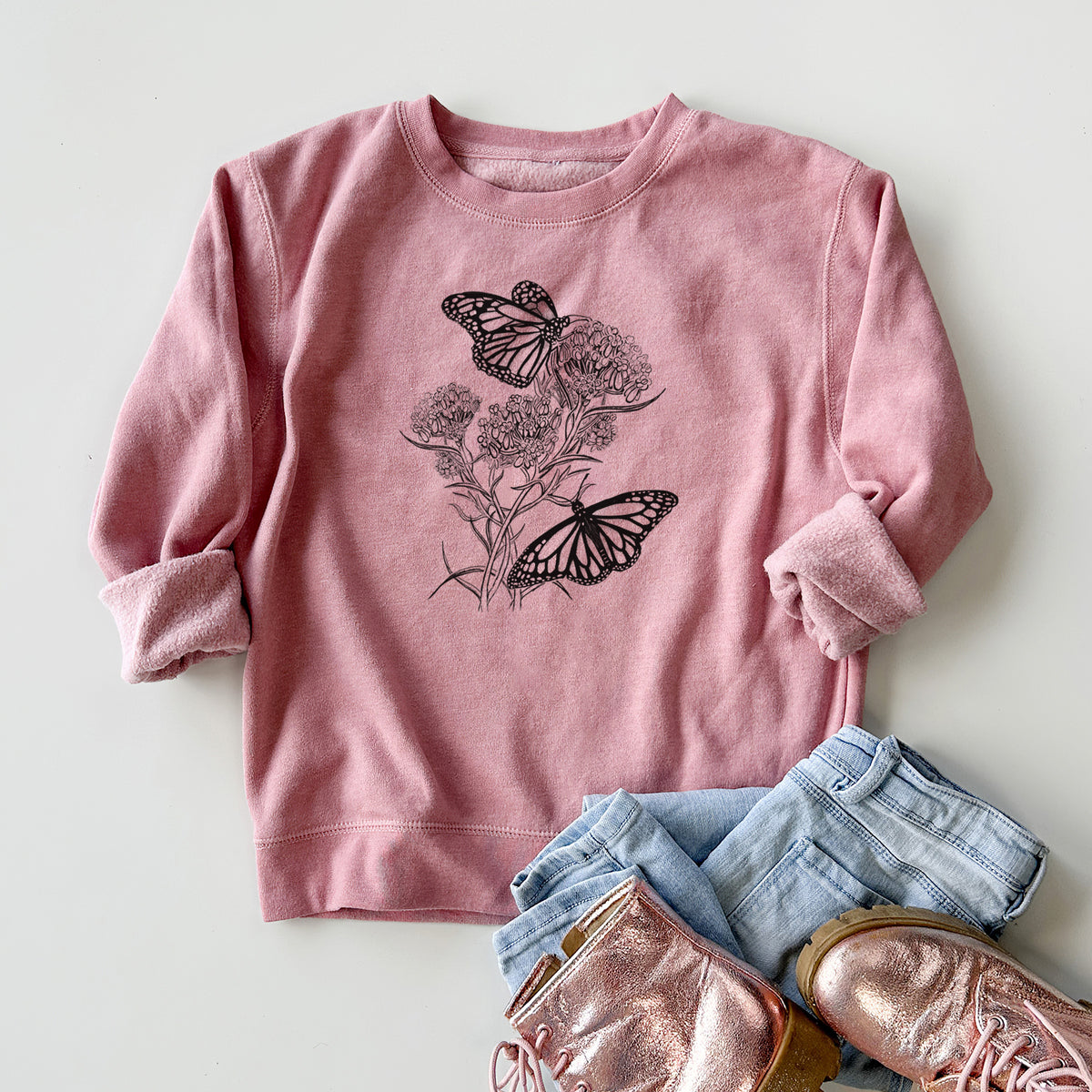 Youth crewneck sweatshirt printed with a black ink illustration of two monarch butterflies on flowering milkweed, styled flat with jeans and boots.