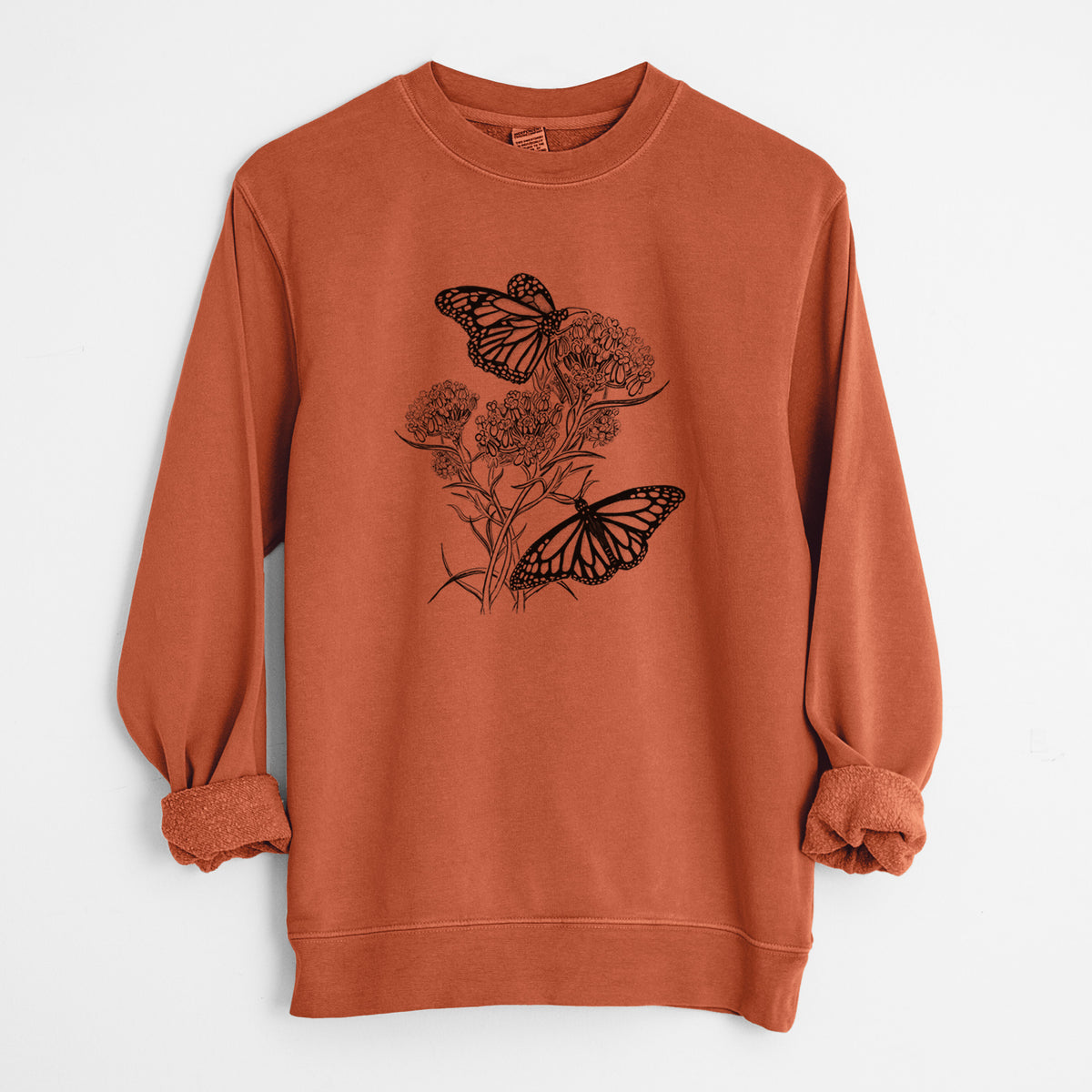 Amber orange pigment dyed crewneck sweatshirt laid flat with the sleeves rolled, printed with a black ink illustration of two monarch butterflies on flowering milkweed.