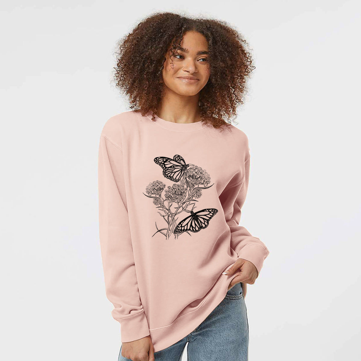 Model wearing a dusty pink pigment dyed crewneck sweatshirt printed with a black ink illustration of two monarch butterflies on flowering milkweed.