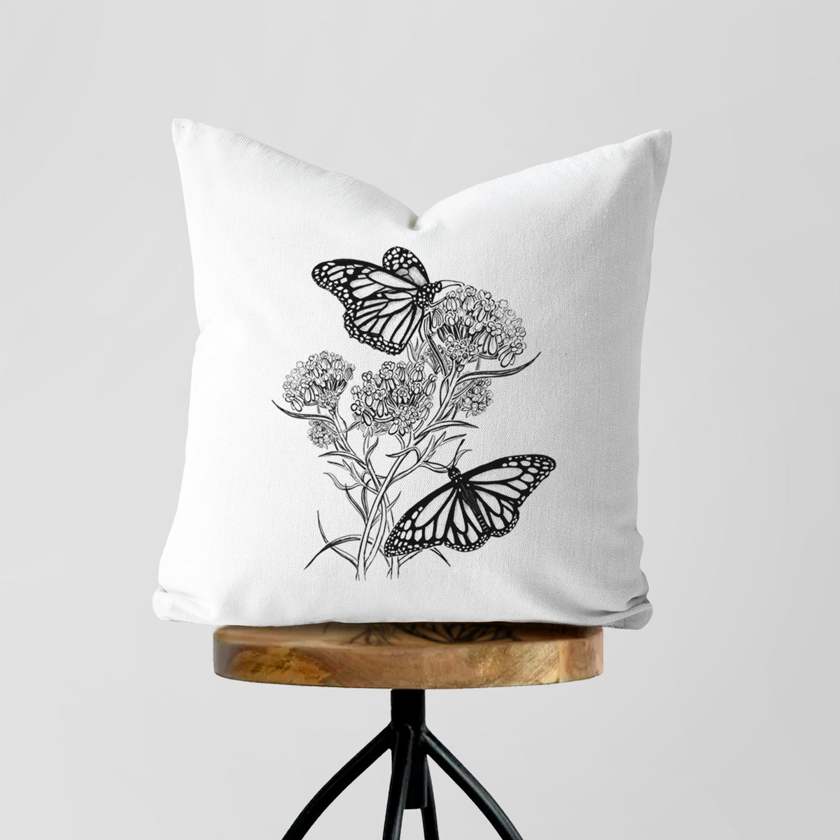 White pillow cover printed with a black ink illustration of two monarch butterflies on flowering milkweed, on a wooden stool.