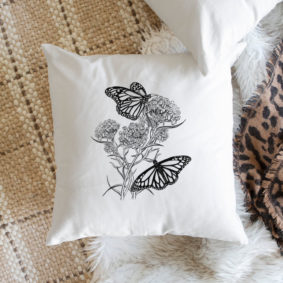 White pillow cover printed with a black ink illustration of two monarch butterflies on flowering milkweed, on a woven chair.