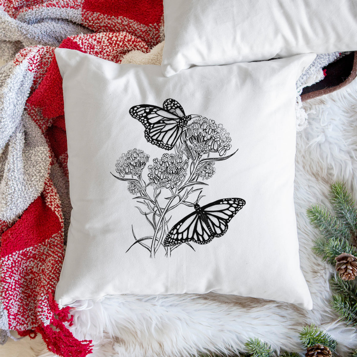 White pillow cover printed with a black ink illustration of two monarch butterflies on flowering milkweed, on a woven chair.