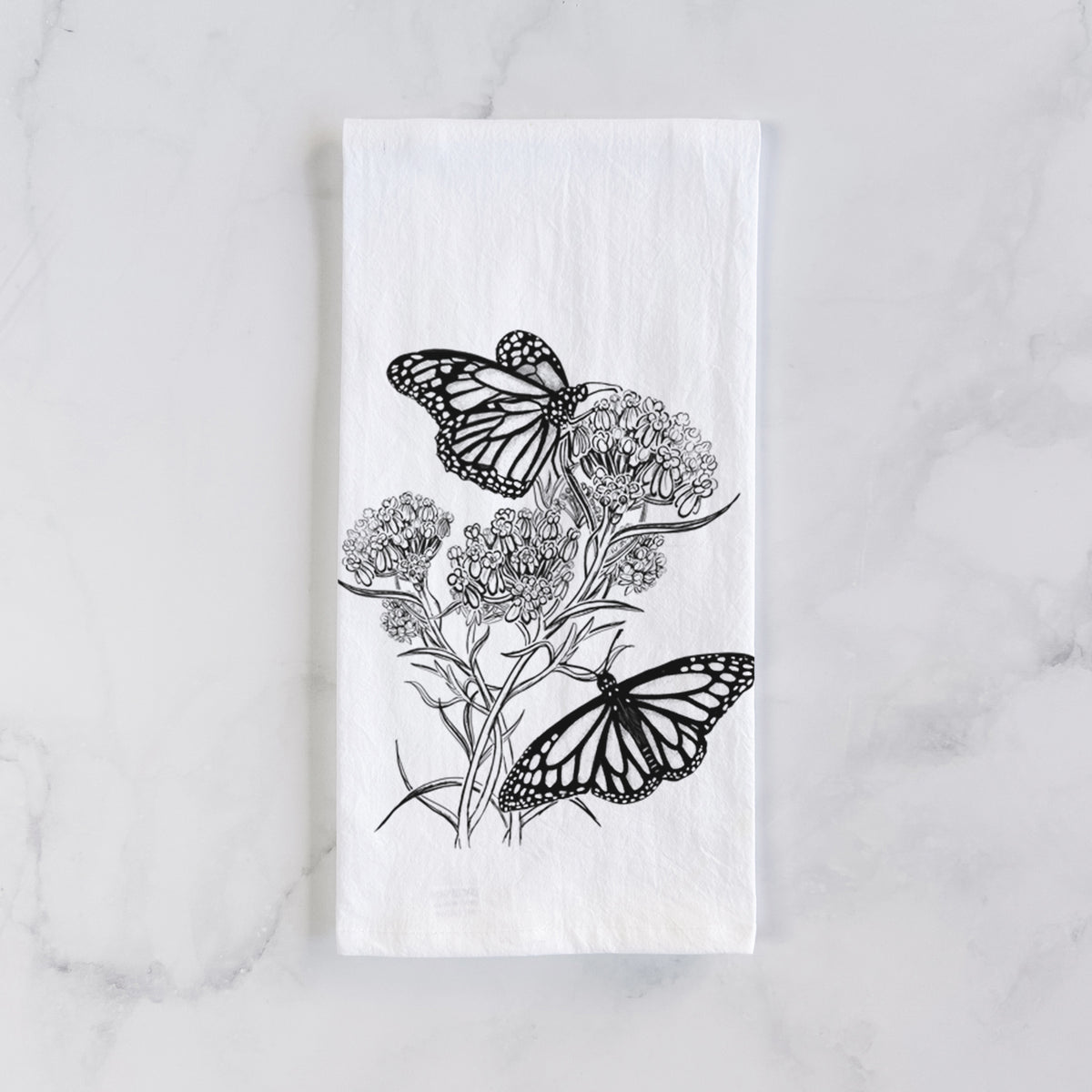 White cotton tea towel laid flat on marble, printed with a black ink illustration of two monarch butterflies on flowering milkweed.