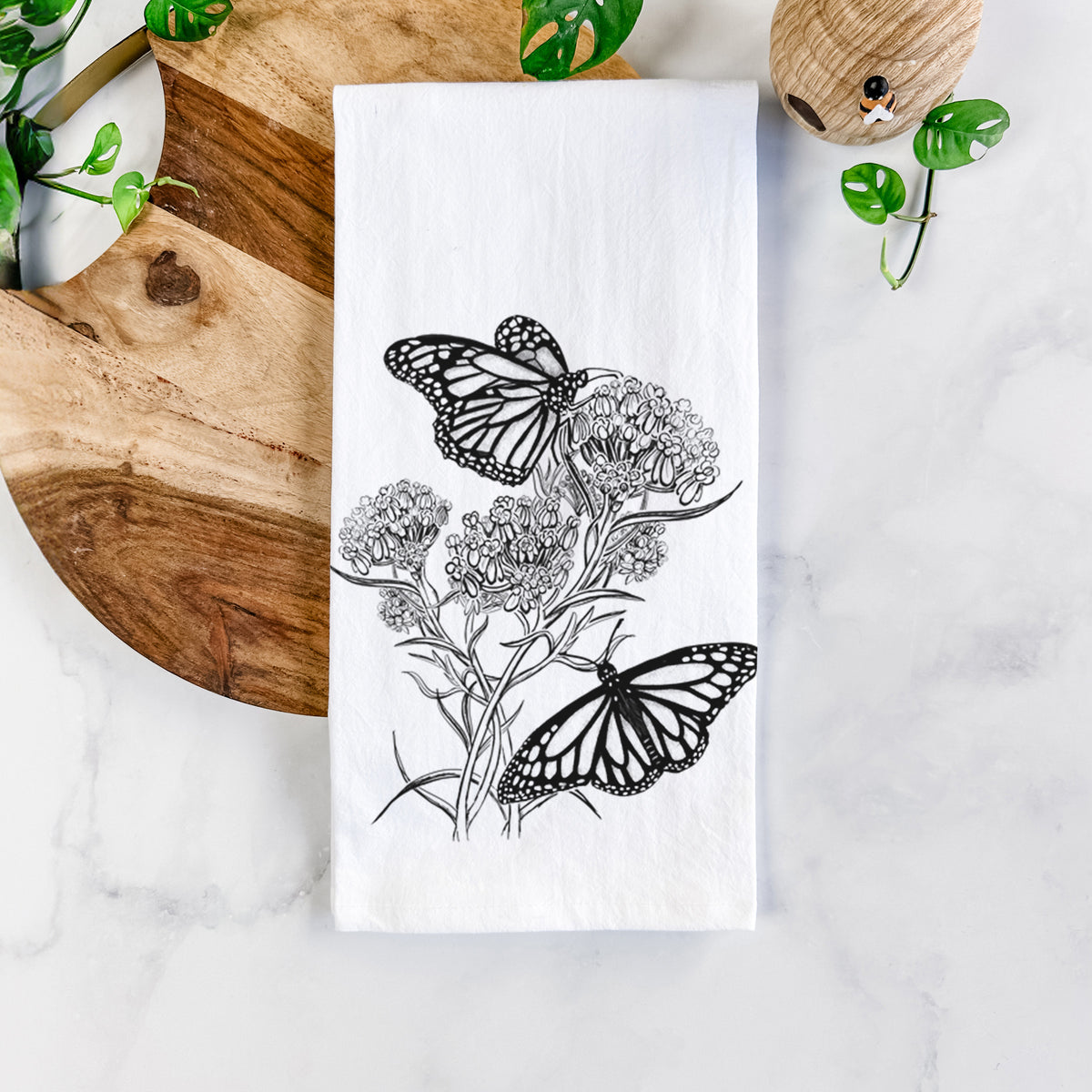 White cotton tea towel printed with a black ink illustration of two monarch butterflies on flowering milkweed, on a marble counter beside a round wooden cutting board and greenery.