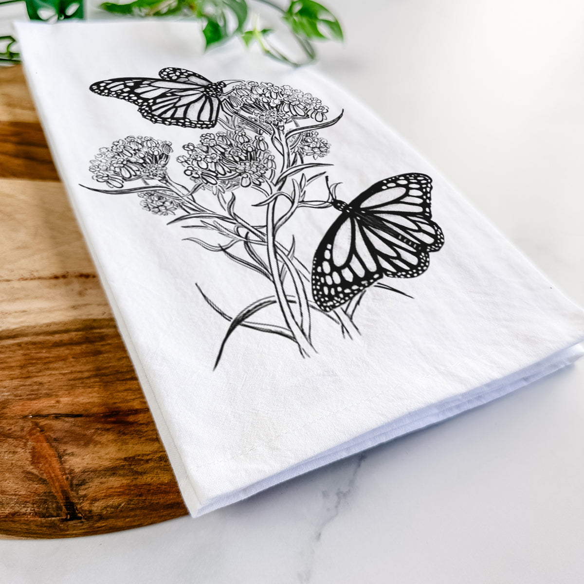 Close-up of a folded white tea towel on a wooden board, showing a black ink illustration of two monarch butterflies on flowering milkweed.