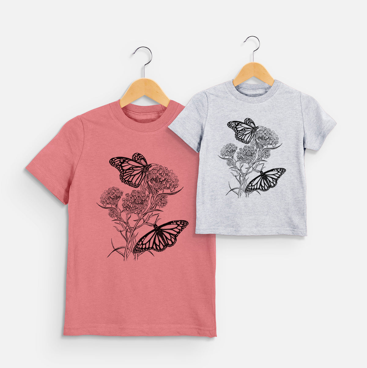 Two youth t-shirts in different colors on hangers, printed with a black ink illustration of two monarch butterflies on flowering milkweed.