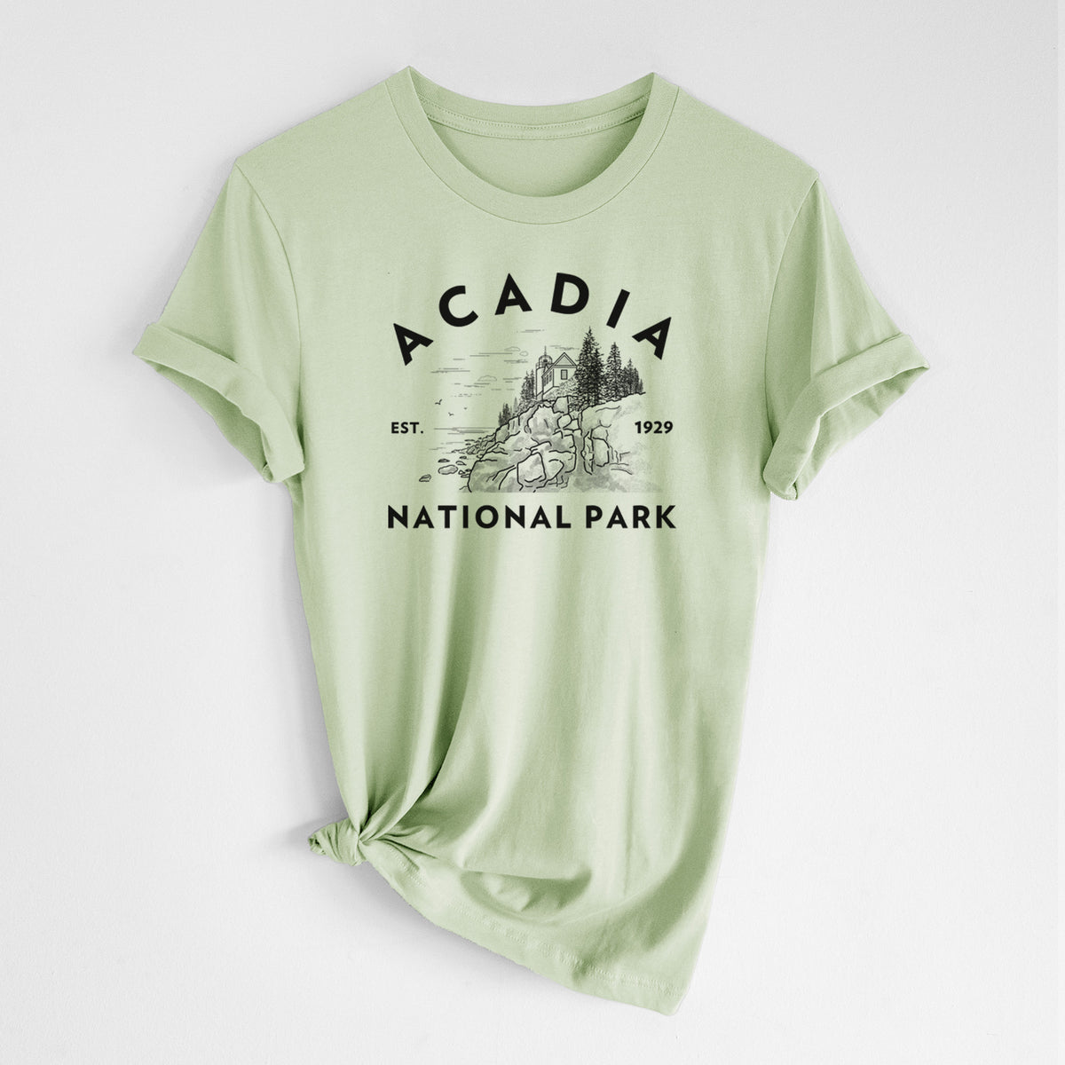 Spring green lightweight cotton t-shirt laid flat with the sleeves rolled, printed with a round badge of a rocky coastline and pine trees, with the words Acadia National Park, Est. 1929.