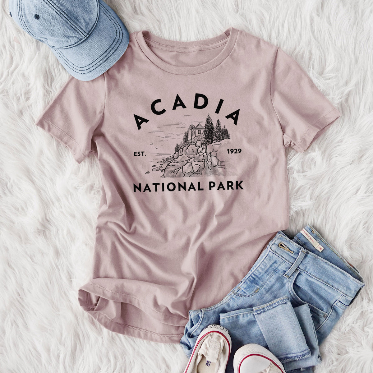 Women's relaxed fit cotton t-shirt printed with a round badge of a rocky coastline and pine trees, with the words Acadia National Park, Est. 1929, styled flat on a white faux fur rug with jeans.