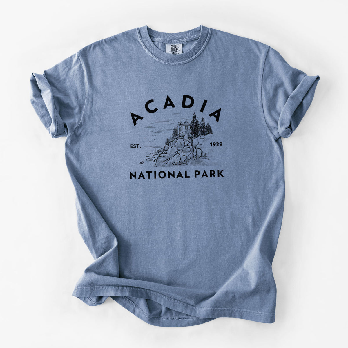 Blue jean Comfort Colors t-shirt laid flat, printed with a round badge of a rocky coastline and pine trees, with the words Acadia National Park, Est. 1929.
