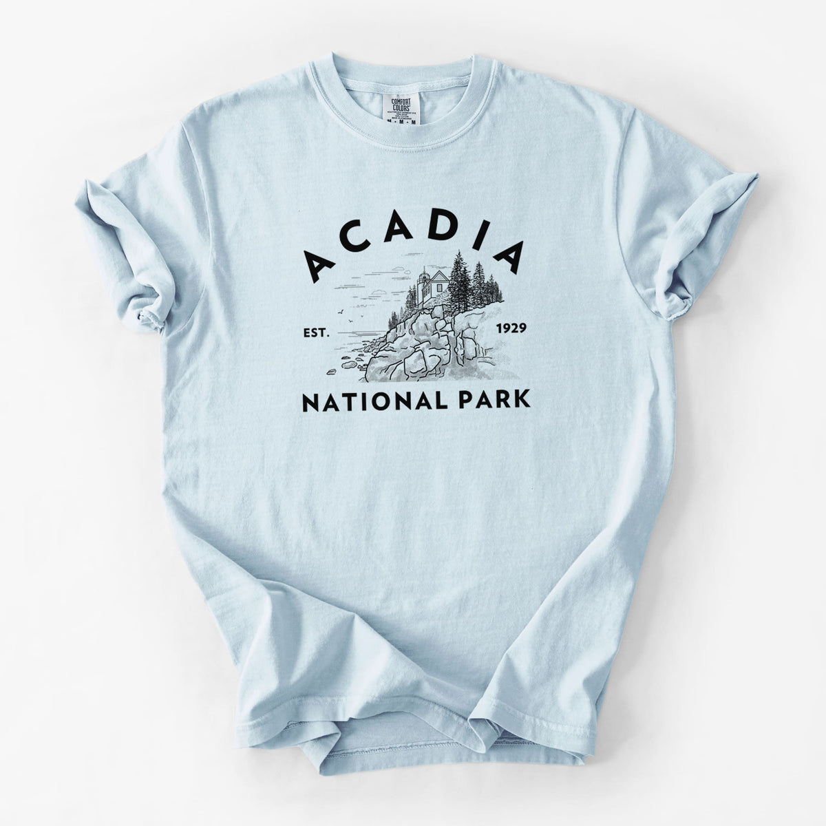 Chambray blue Comfort Colors t-shirt laid flat, printed with a round badge of a rocky coastline and pine trees, with the words Acadia National Park, Est. 1929.