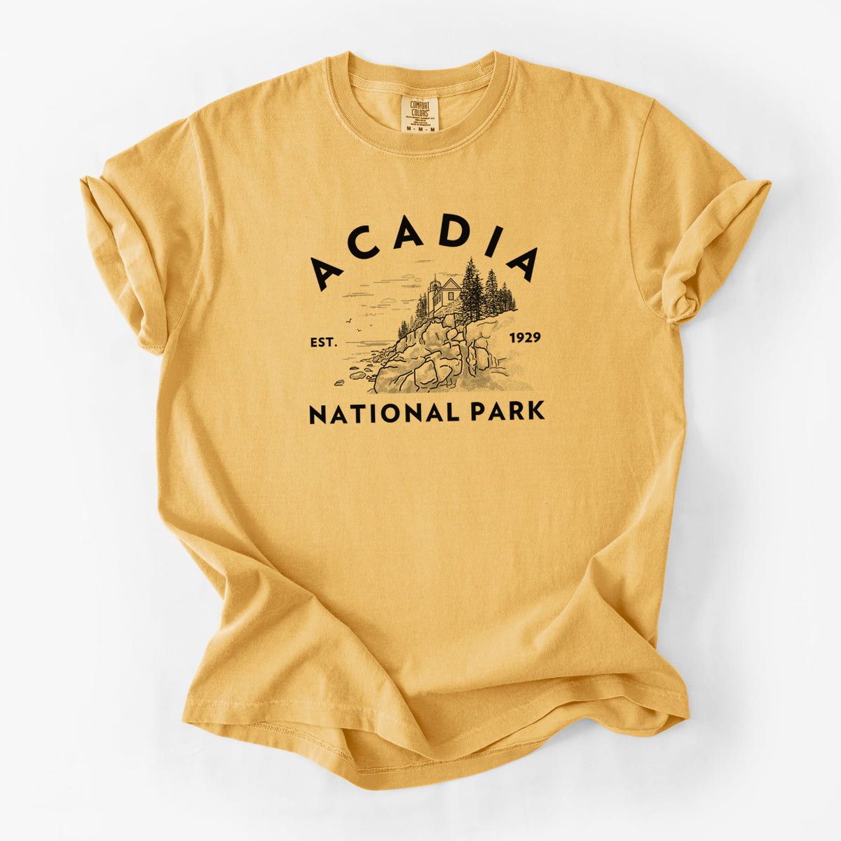 Golden wheat yellow Comfort Colors t-shirt laid flat, printed with a round badge of a rocky coastline and pine trees, with the words Acadia National Park, Est. 1929.