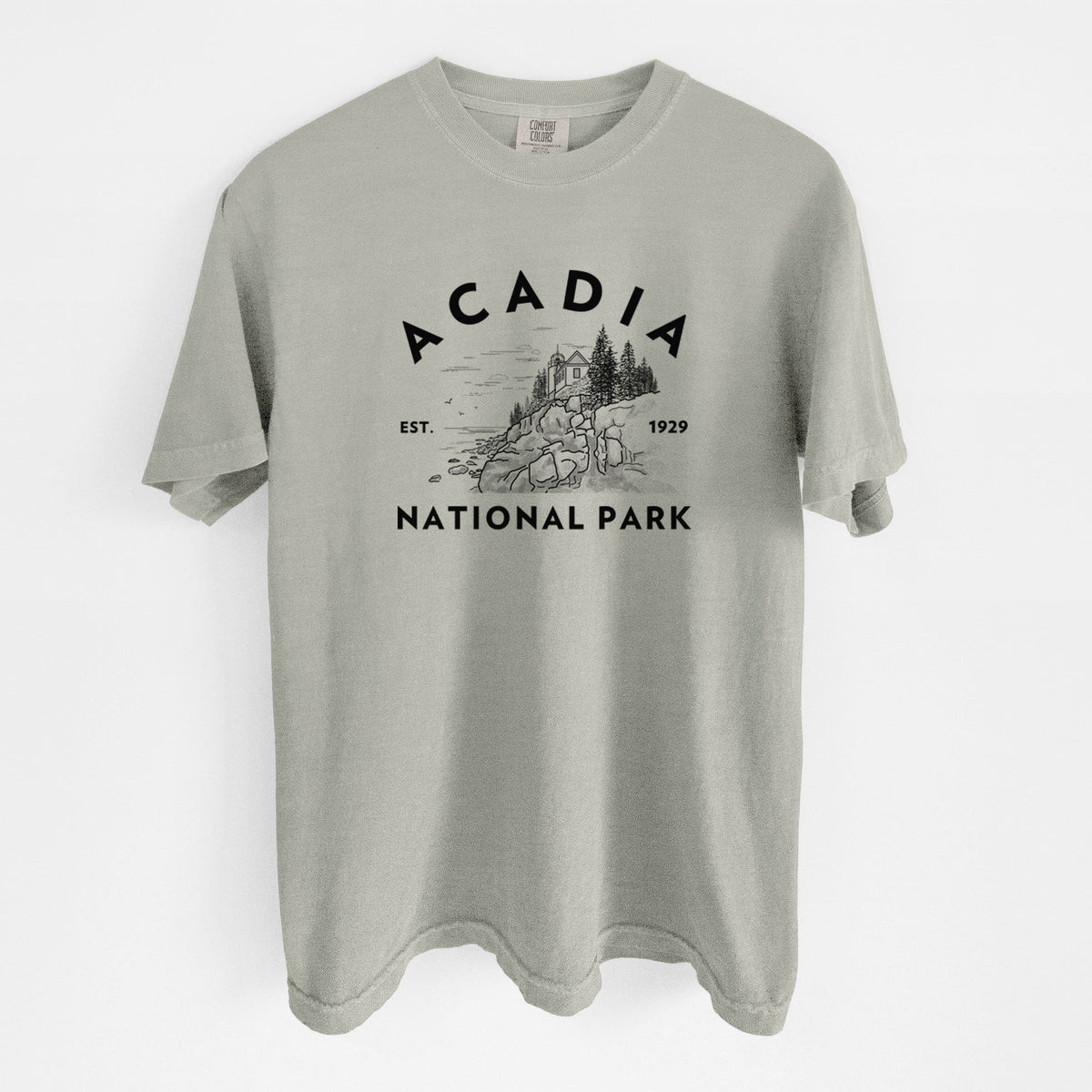 Sandstone beige Comfort Colors t-shirt laid flat, printed with a round badge of a rocky coastline and pine trees, with the words Acadia National Park, Est. 1929.