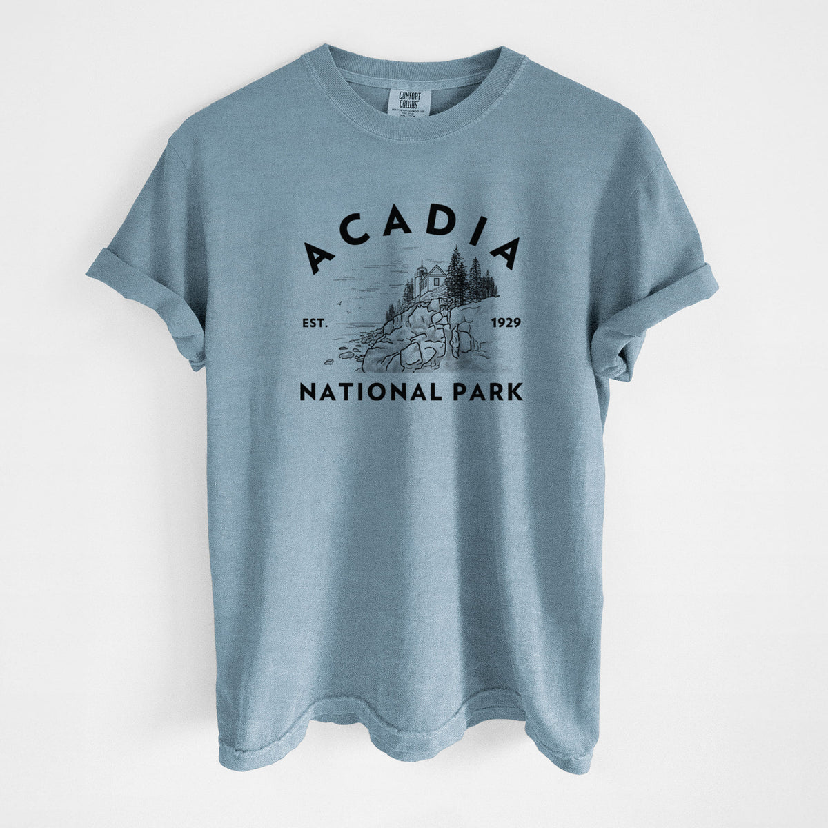 Steel blue Comfort Colors t-shirt laid flat, printed with a round badge of a rocky coastline and pine trees, with the words Acadia National Park, Est. 1929.