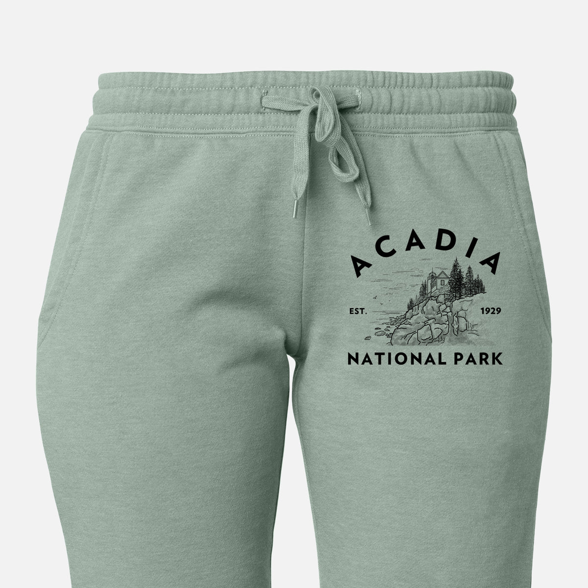 Front of sage green Cali Wave sweatpants, printed with a round badge of a rocky coastline and pine trees, with the words Acadia National Park, Est. 1929 on the leg.