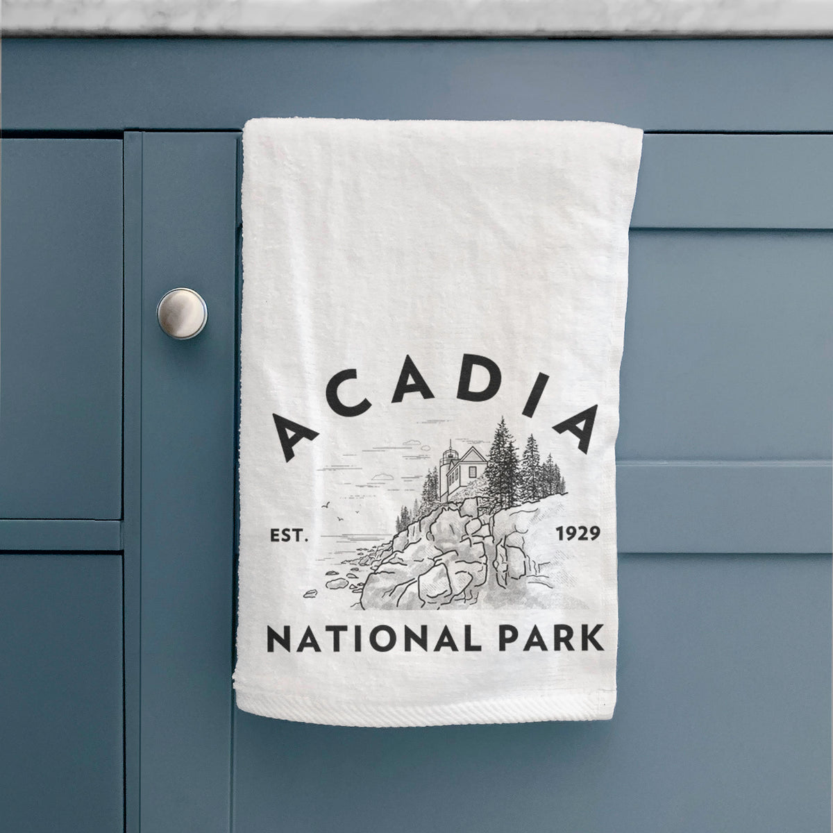 White hand towel printed with a round badge of a rocky coastline and pine trees, with the words Acadia National Park, Est. 1929, hanging on a cabinet door.