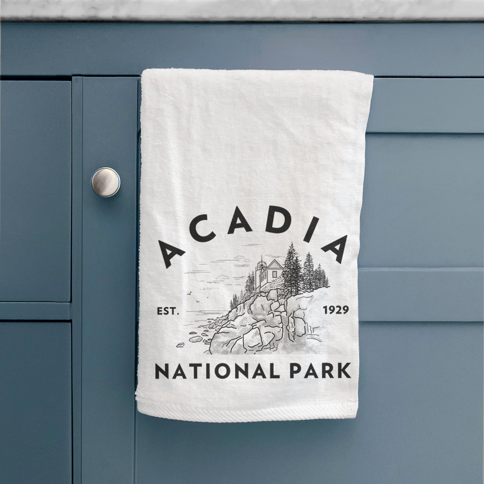 White hand towel printed with a round badge of a rocky coastline and pine trees, with the words Acadia National Park, Est. 1929.