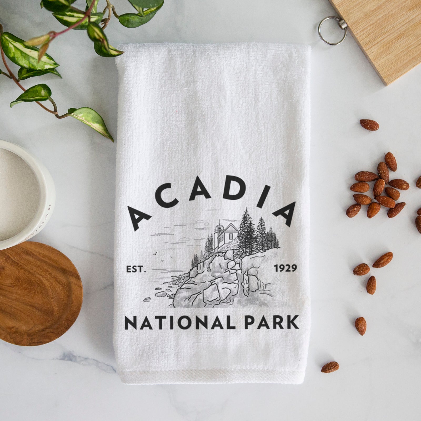 White hand towel printed with a round badge of a rocky coastline and pine trees, with the words Acadia National Park, Est. 1929.