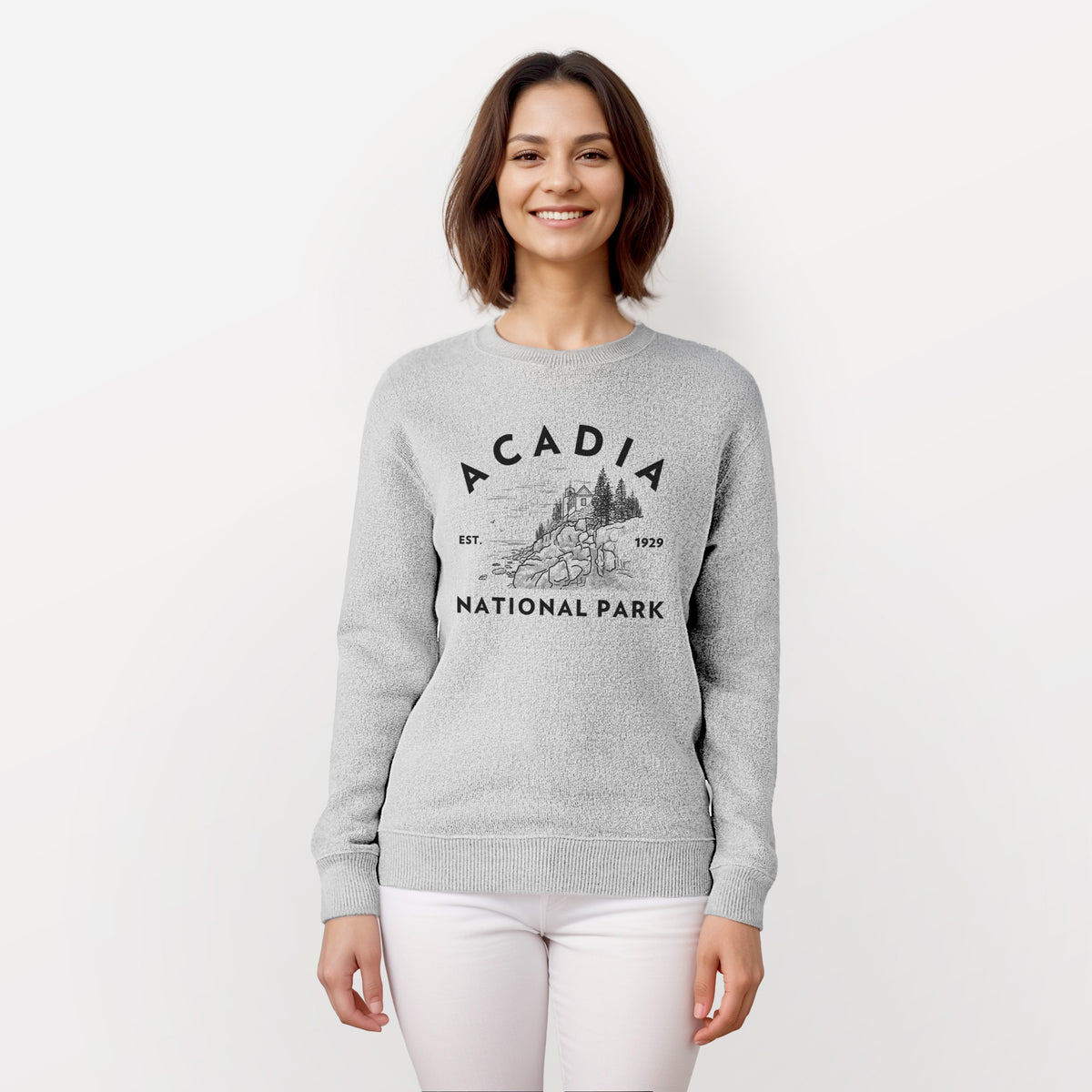 Woman wearing a knit sweatshirt printed with a round badge of a rocky coastline and pine trees, with the words Acadia National Park, Est. 1929.