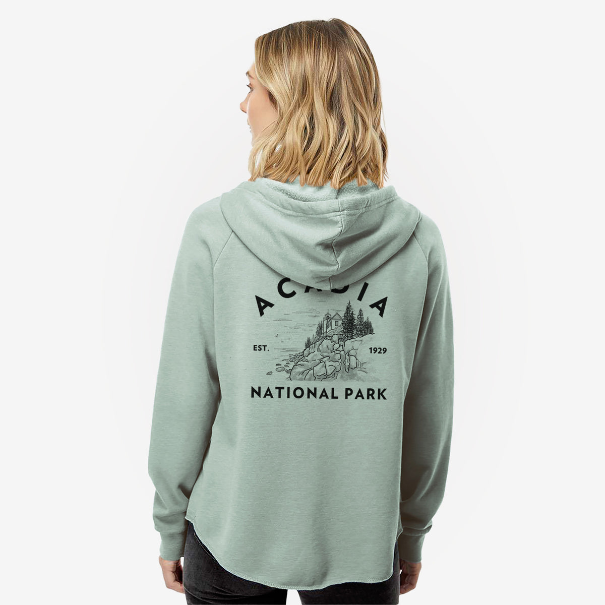 Woman wearing a sage green women's Cali Wave zip-up sweatshirt printed with a round badge of a rocky coastline and pine trees, with the words Acadia National Park, Est. 1929, seen from behind.