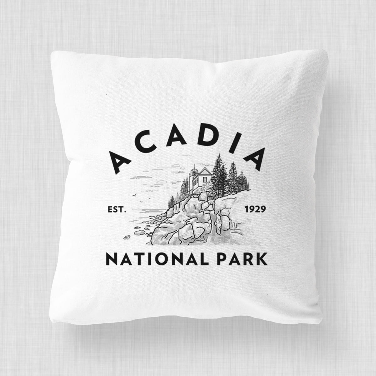 White pillow cover printed with a round badge of a rocky coastline and pine trees, with the words Acadia National Park, Est. 1929.