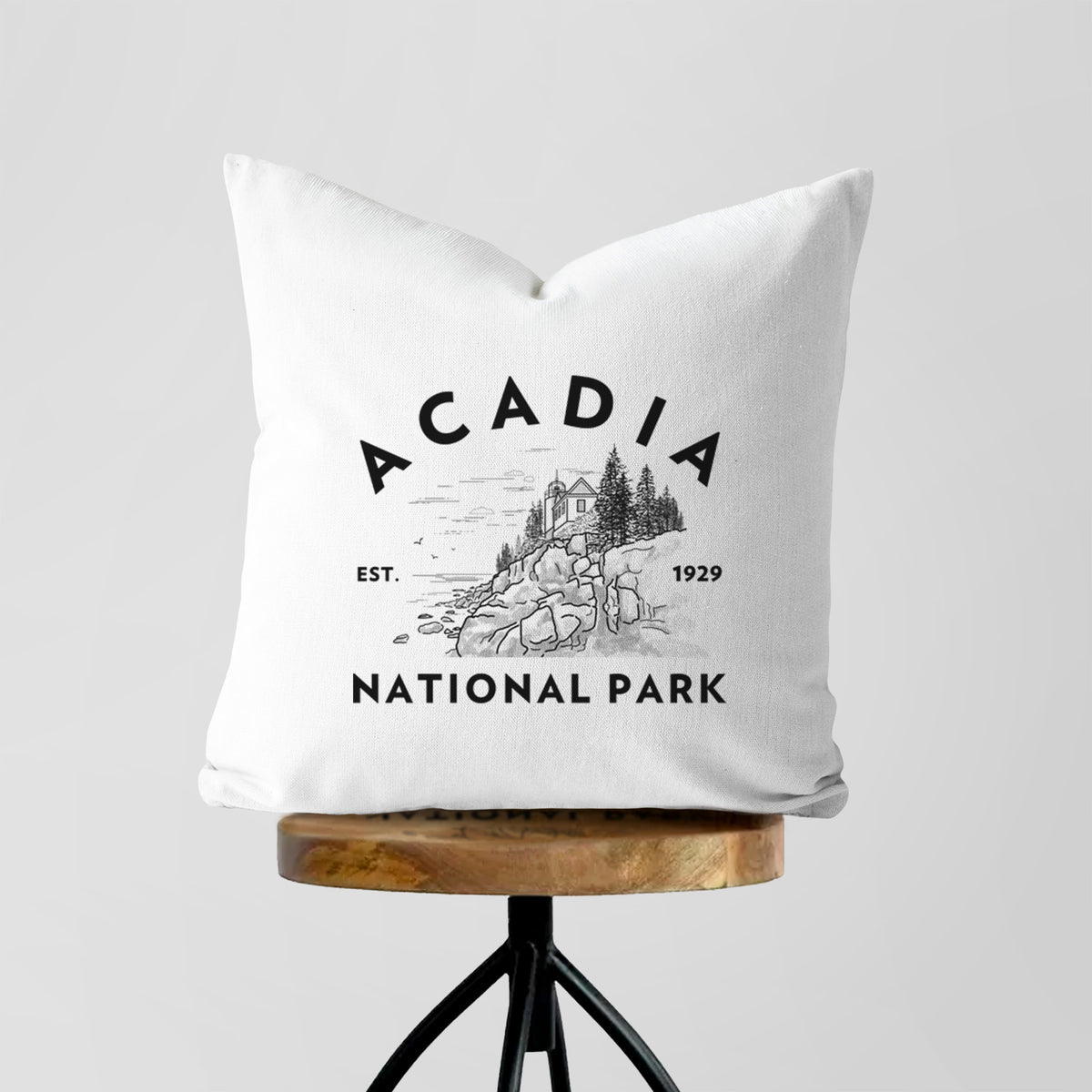 White pillow cover printed with a round badge of a rocky coastline and pine trees, with the words Acadia National Park, Est. 1929, on a wooden stool.