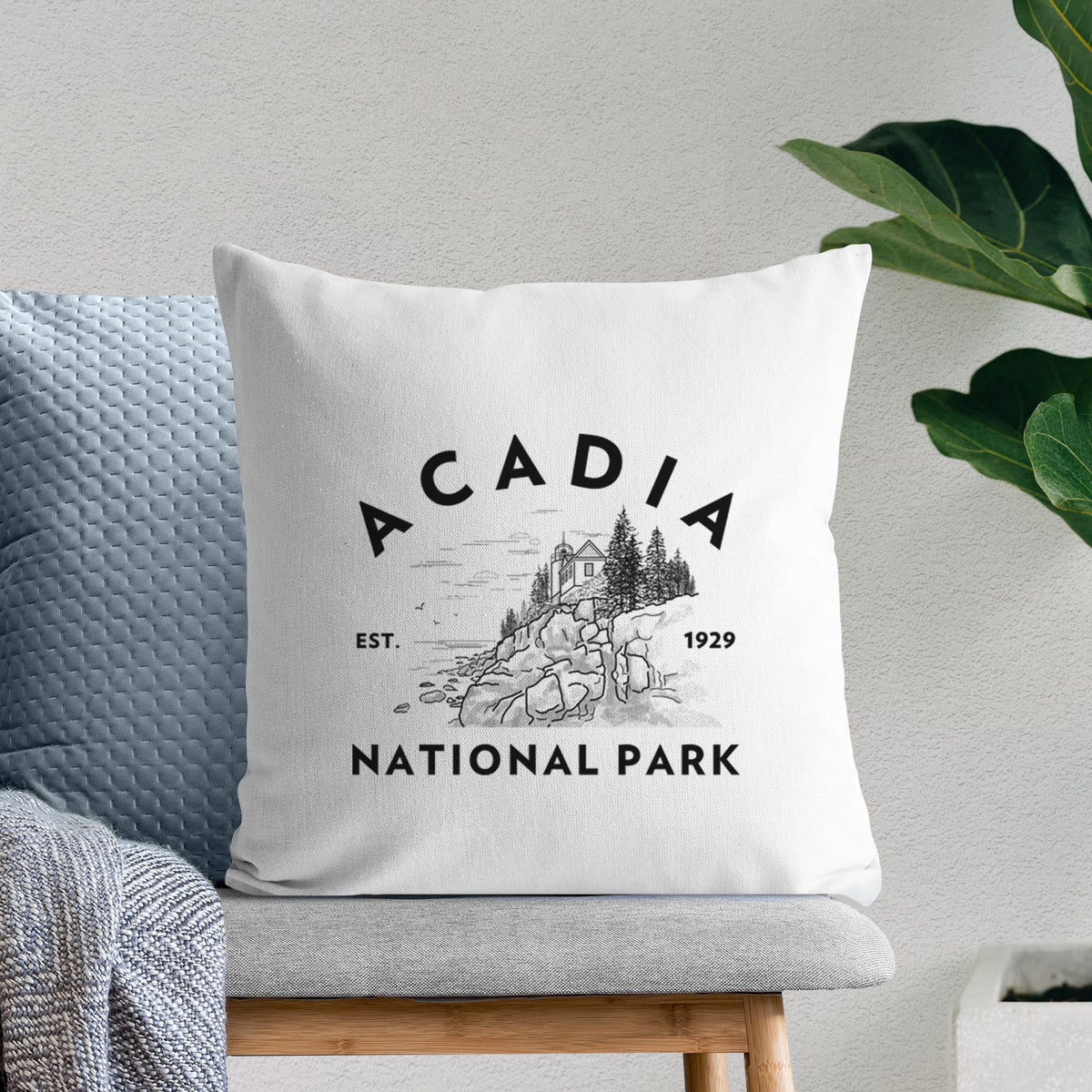 White pillow cover printed with a round badge of a rocky coastline and pine trees, with the words Acadia National Park, Est. 1929, on a bench beside plants.