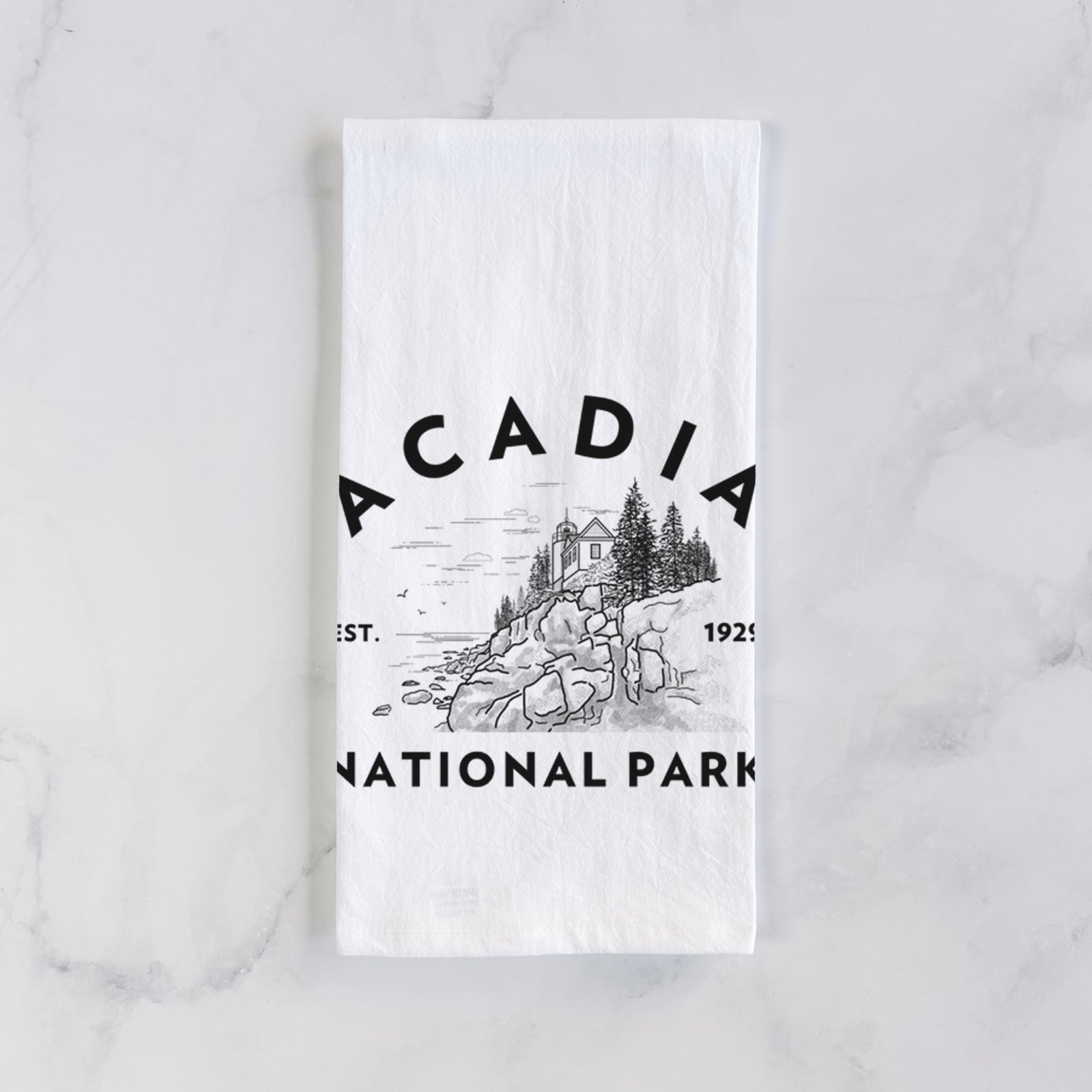 White cotton tea towel printed with a round badge of a rocky coastline and pine trees, with the words Acadia National Park, Est. 1929, on a marble counter beside a round wooden cutting board and greenery.