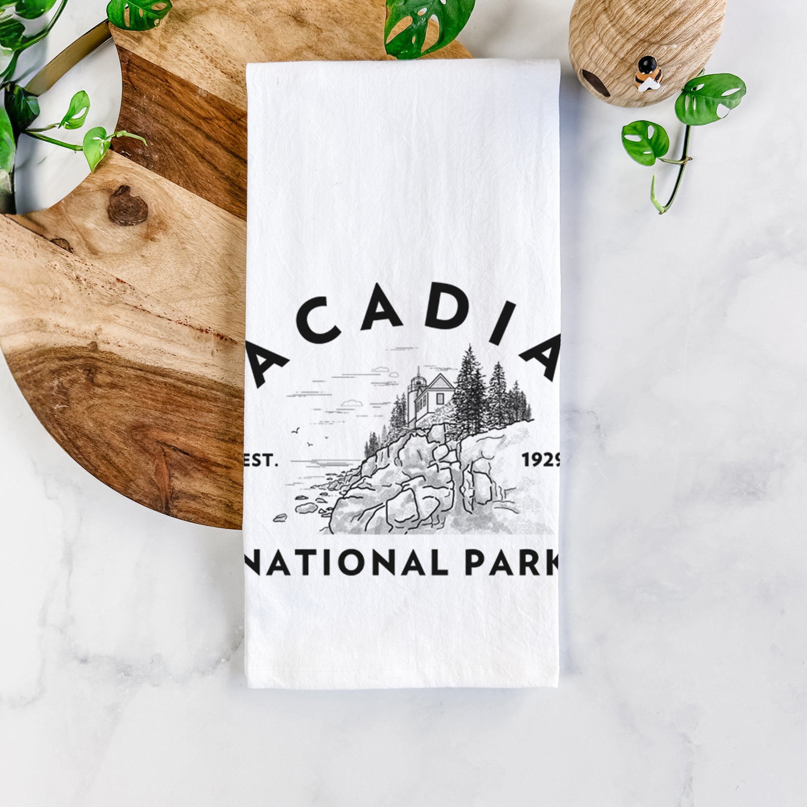 White cotton tea towel printed with a round badge of a rocky coastline and pine trees, with the words Acadia National Park, Est. 1929, on a marble counter beside a round wooden cutting board and greenery.