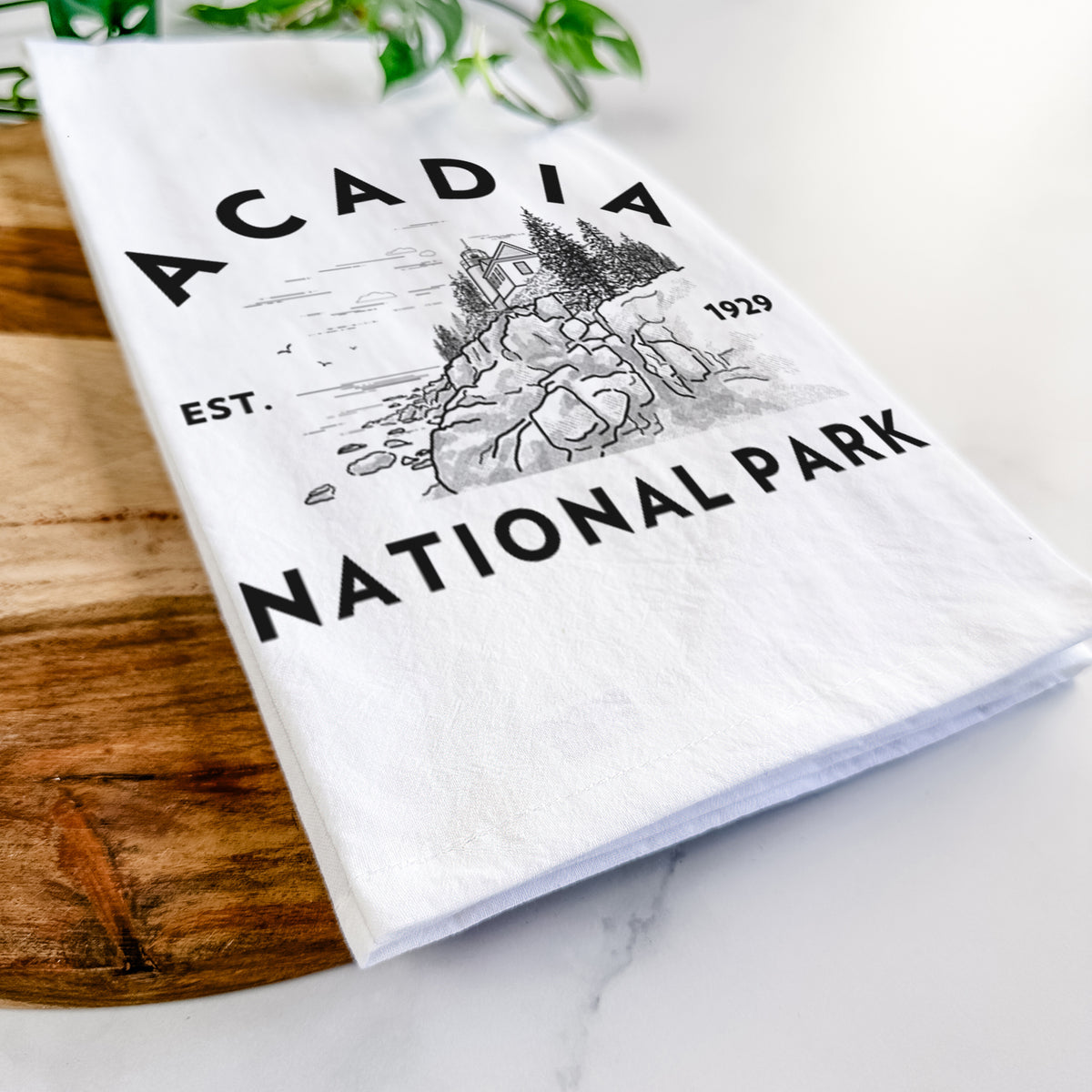 Close-up of a folded white tea towel on a wooden board, showing a round badge of a rocky coastline and pine trees, with the words Acadia National Park, Est. 1929.