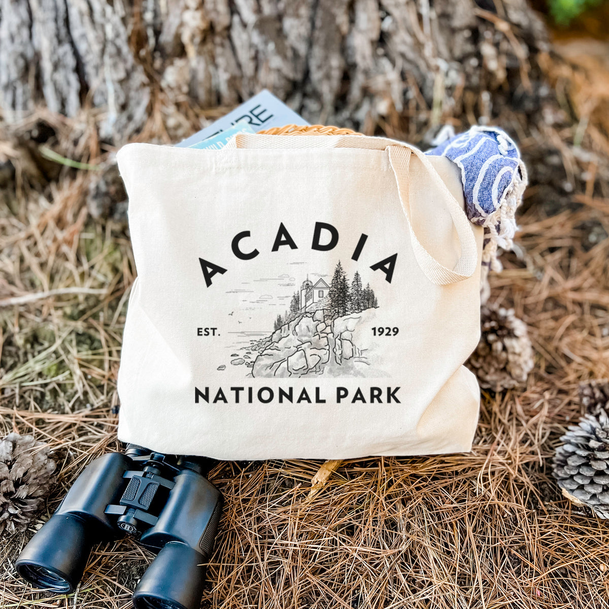 Canvas tote bag printed with a round badge of a rocky coastline and pine trees, with the words Acadia National Park, Est. 1929, resting on dry grass beside a pair of binoculars.
