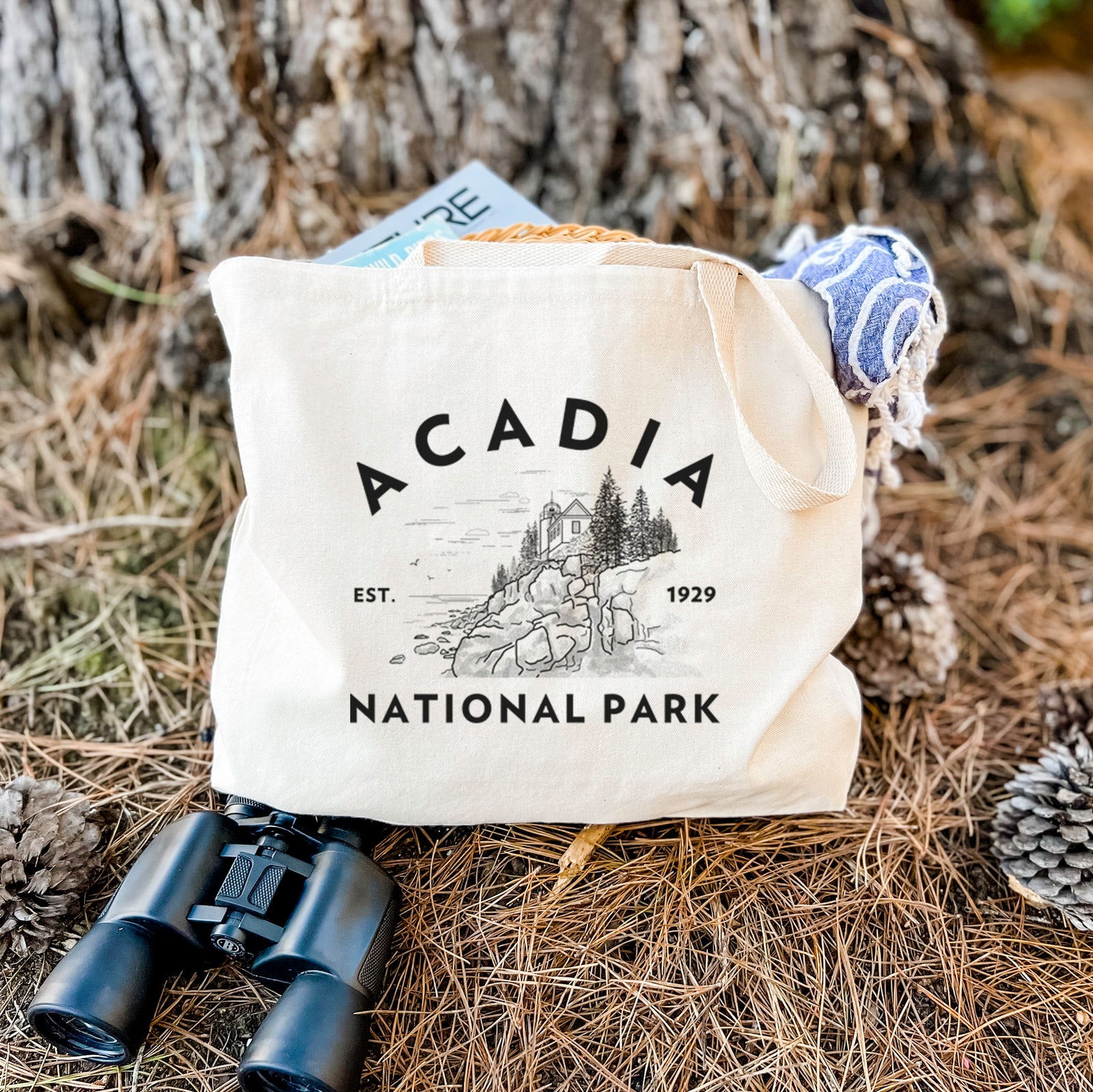 Natural canvas tote bag printed with a round badge of a rocky coastline and pine trees, with the words Acadia National Park, Est. 1929.