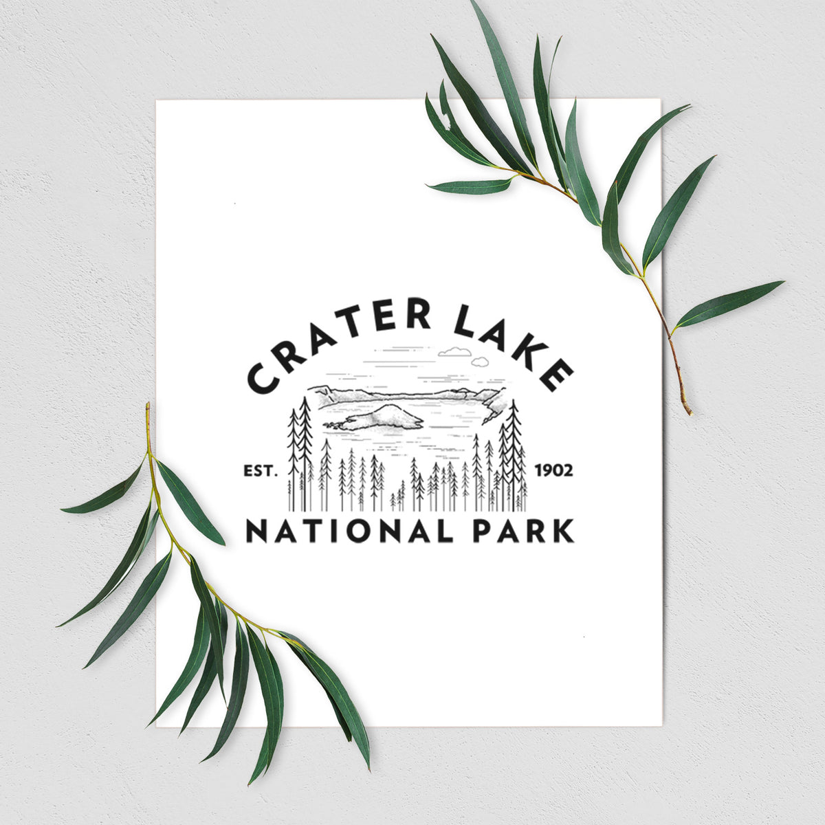 Art print of a round badge of Crater Lake ringed by pine trees, with the words Crater Lake National Park, Est. 1902, shown with eucalyptus leaves.