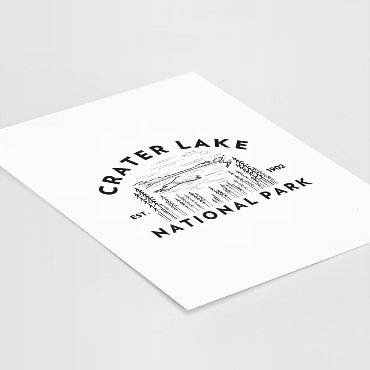 Art print of a round badge of Crater Lake ringed by pine trees, with the words Crater Lake National Park, Est. 1902, lying at an angle on a white surface.