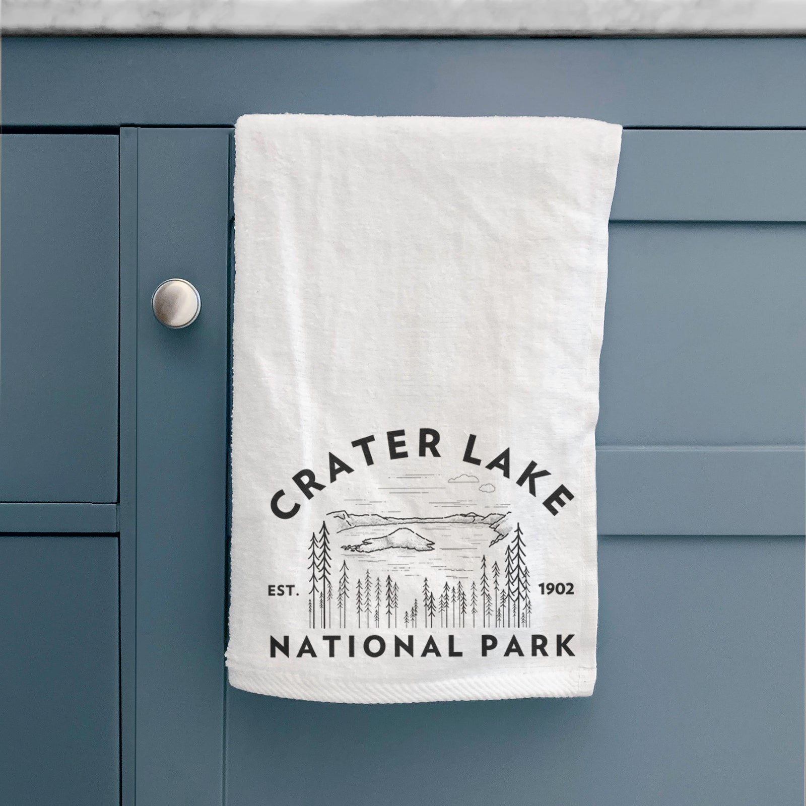 White hand towel printed with a round badge of Crater Lake ringed by pine trees, with the words Crater Lake National Park, Est. 1902.