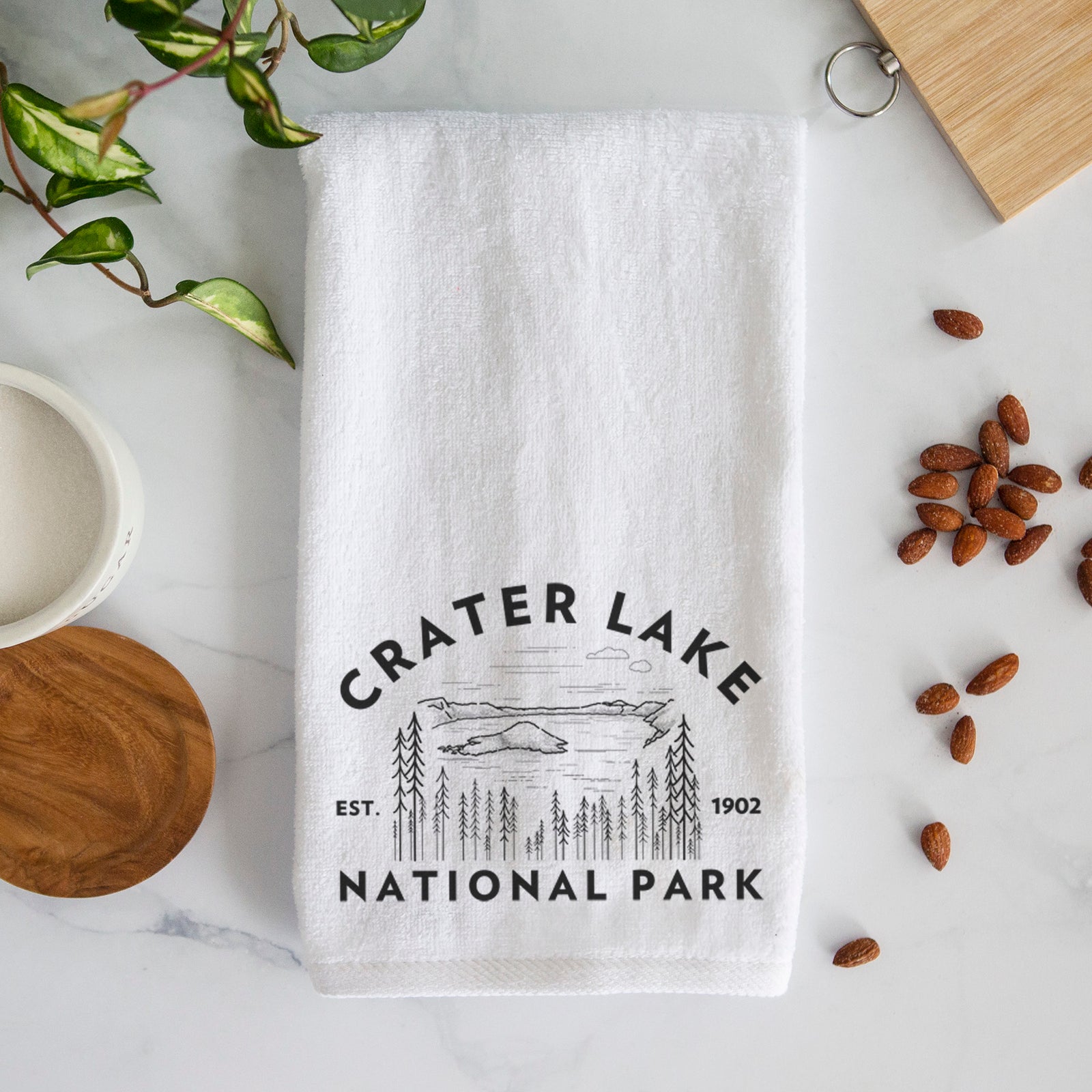 White hand towel printed with a round badge of Crater Lake ringed by pine trees, with the words Crater Lake National Park, Est. 1902.