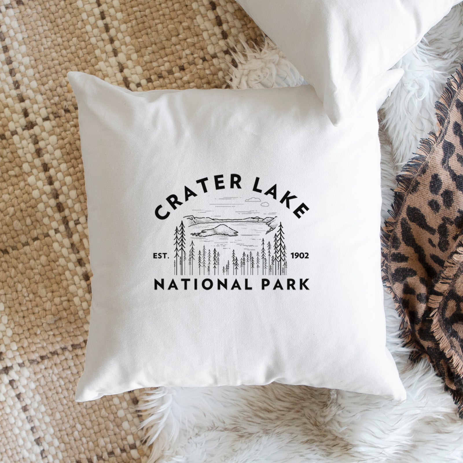 White pillow cover printed with a round badge of Crater Lake ringed by pine trees, with the words Crater Lake National Park, Est. 1902, on a bench beside plants.