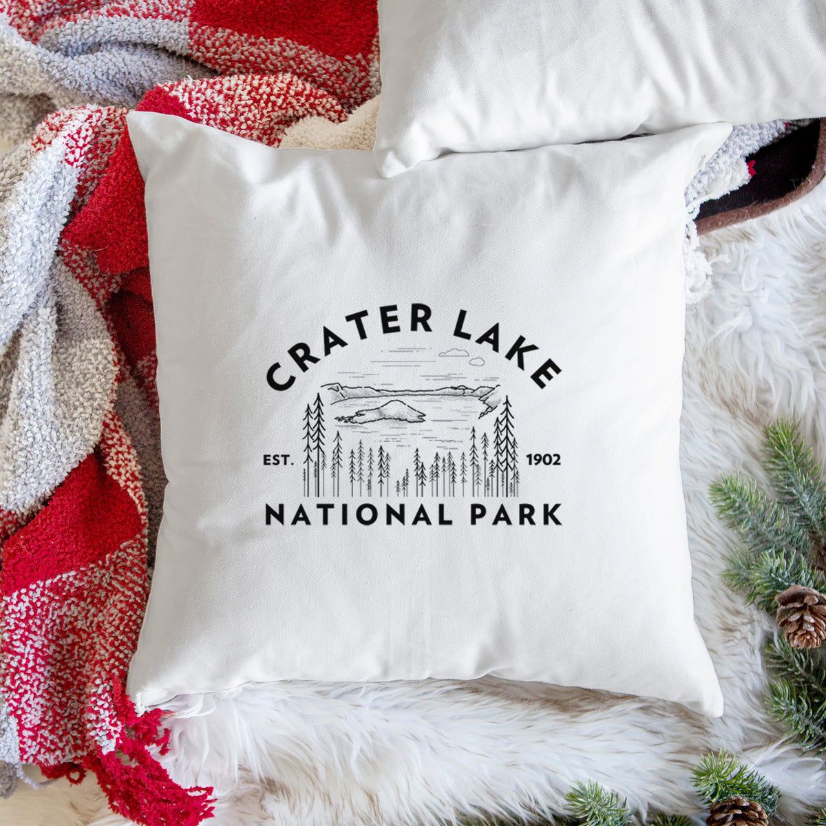 White pillow cover printed with a round badge of Crater Lake ringed by pine trees, with the words Crater Lake National Park, Est. 1902, on a woven chair.