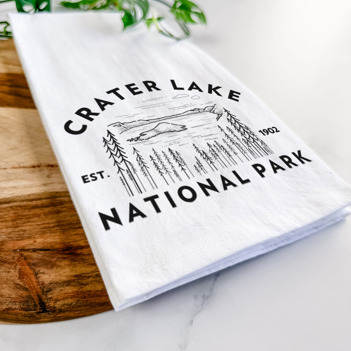 Close-up of a folded white tea towel on a wooden board, showing a round badge of Crater Lake ringed by pine trees, with the words Crater Lake National Park, Est. 1902.