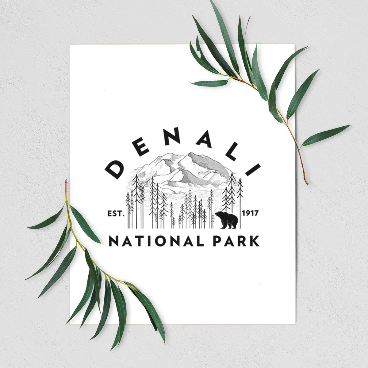 Art print of a round badge of mountains, pine trees and a bear, with the words Denali National Park, Est. 1917, shown with eucalyptus leaves.