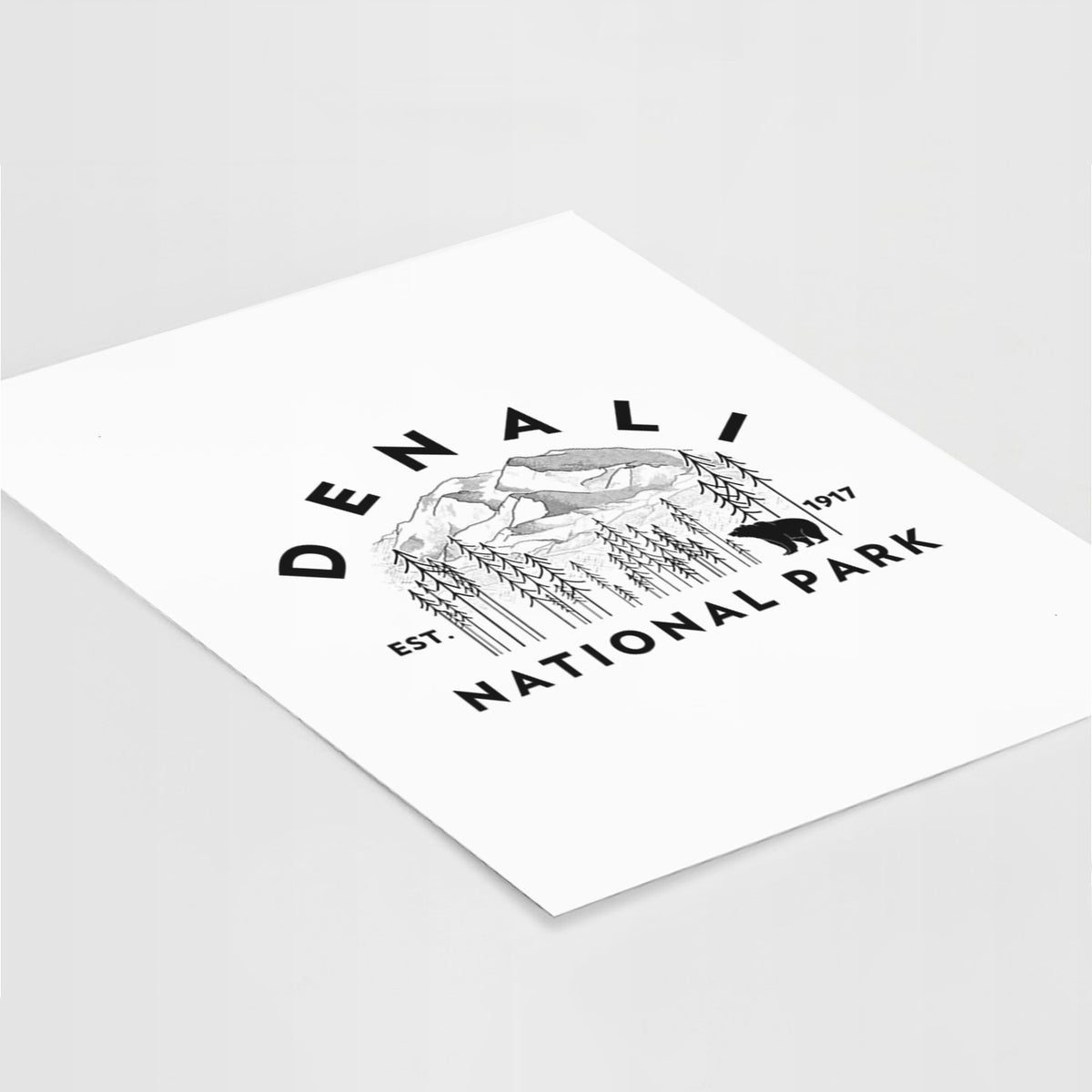 Art print of a round badge of mountains, pine trees and a bear, with the words Denali National Park, Est. 1917, lying at an angle on a white surface.