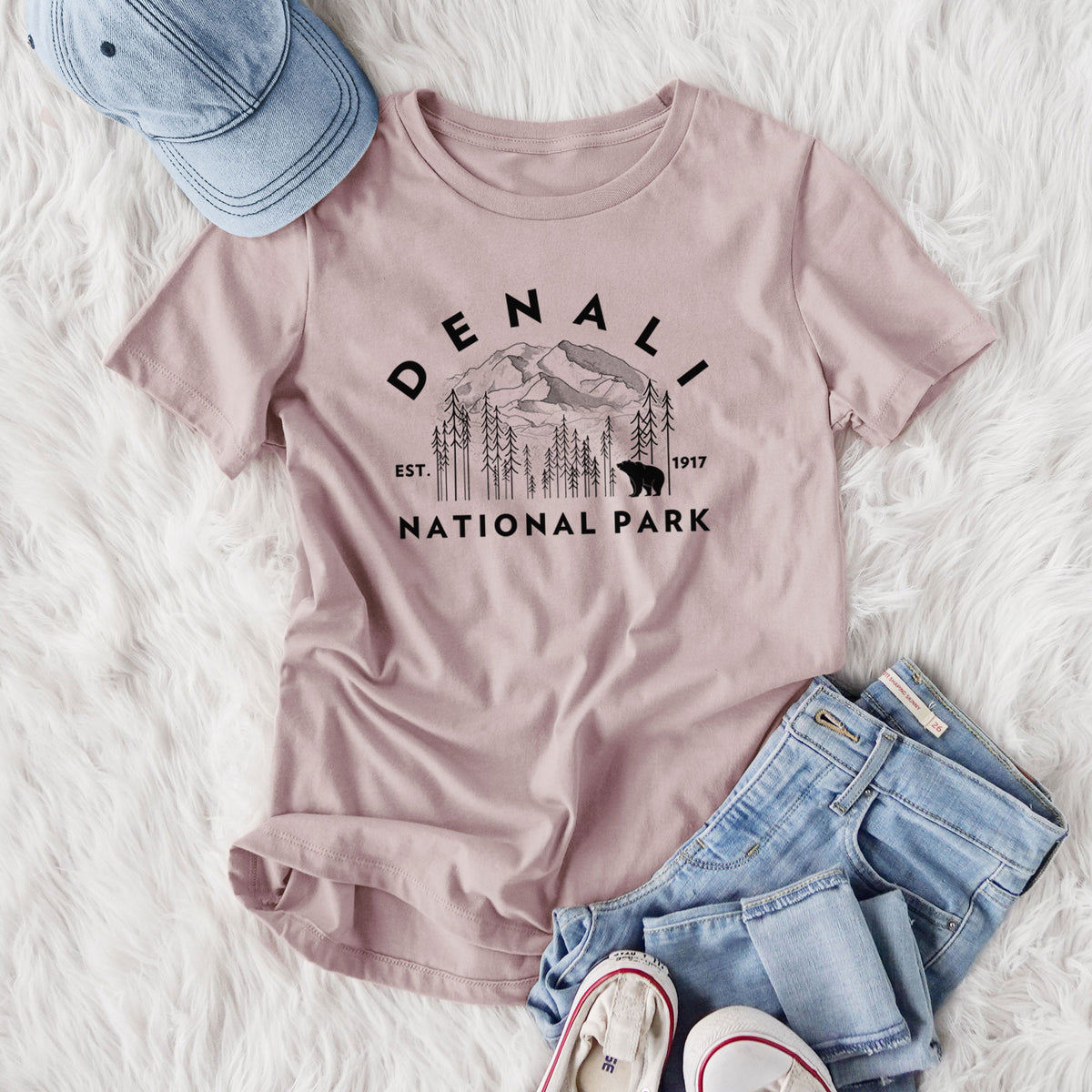 Women's relaxed fit cotton t-shirt printed with a round badge of mountains, pine trees and a bear, with the words Denali National Park, Est. 1917, styled flat on a white faux fur rug with jeans.