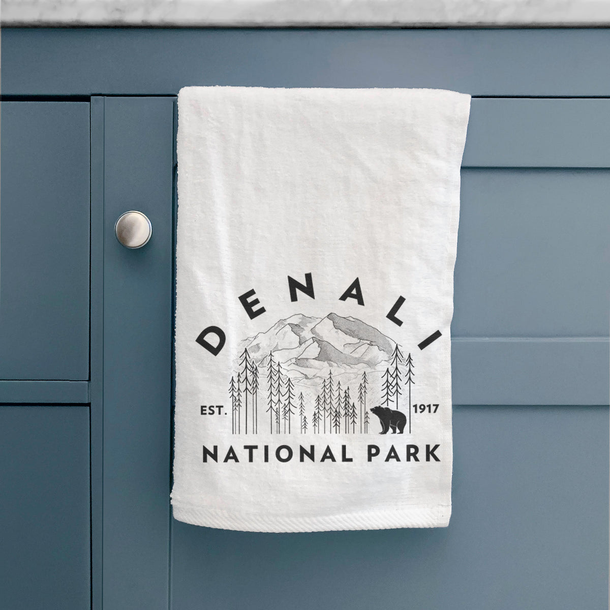 White hand towel printed with a round badge of mountains, pine trees and a bear, with the words Denali National Park, Est. 1917, hanging on a cabinet door.
